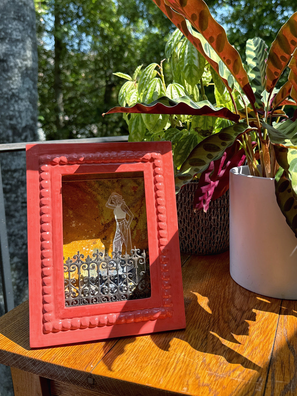 Red framed artwork on a wooden surface with plants in the background