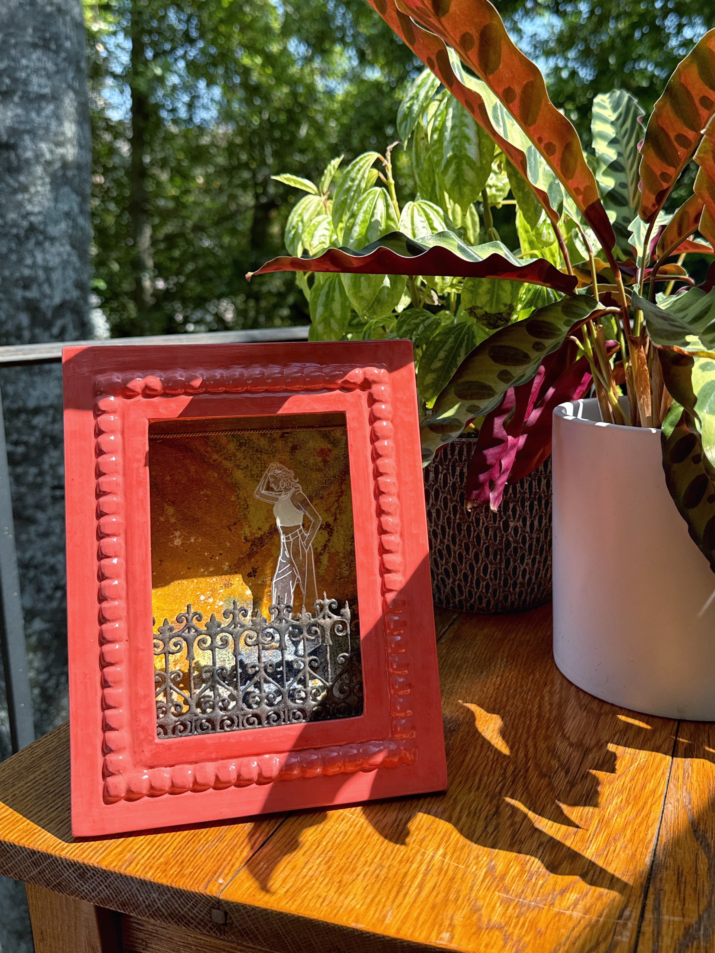 Red framed artwork on a wooden surface with plants in the background
