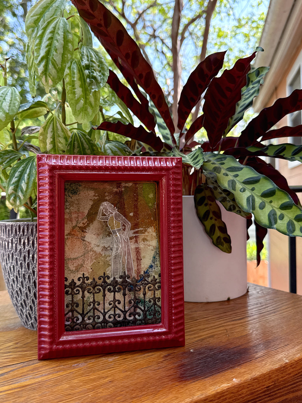 Red framed artwork on a wooden surface with Southern plants in the background