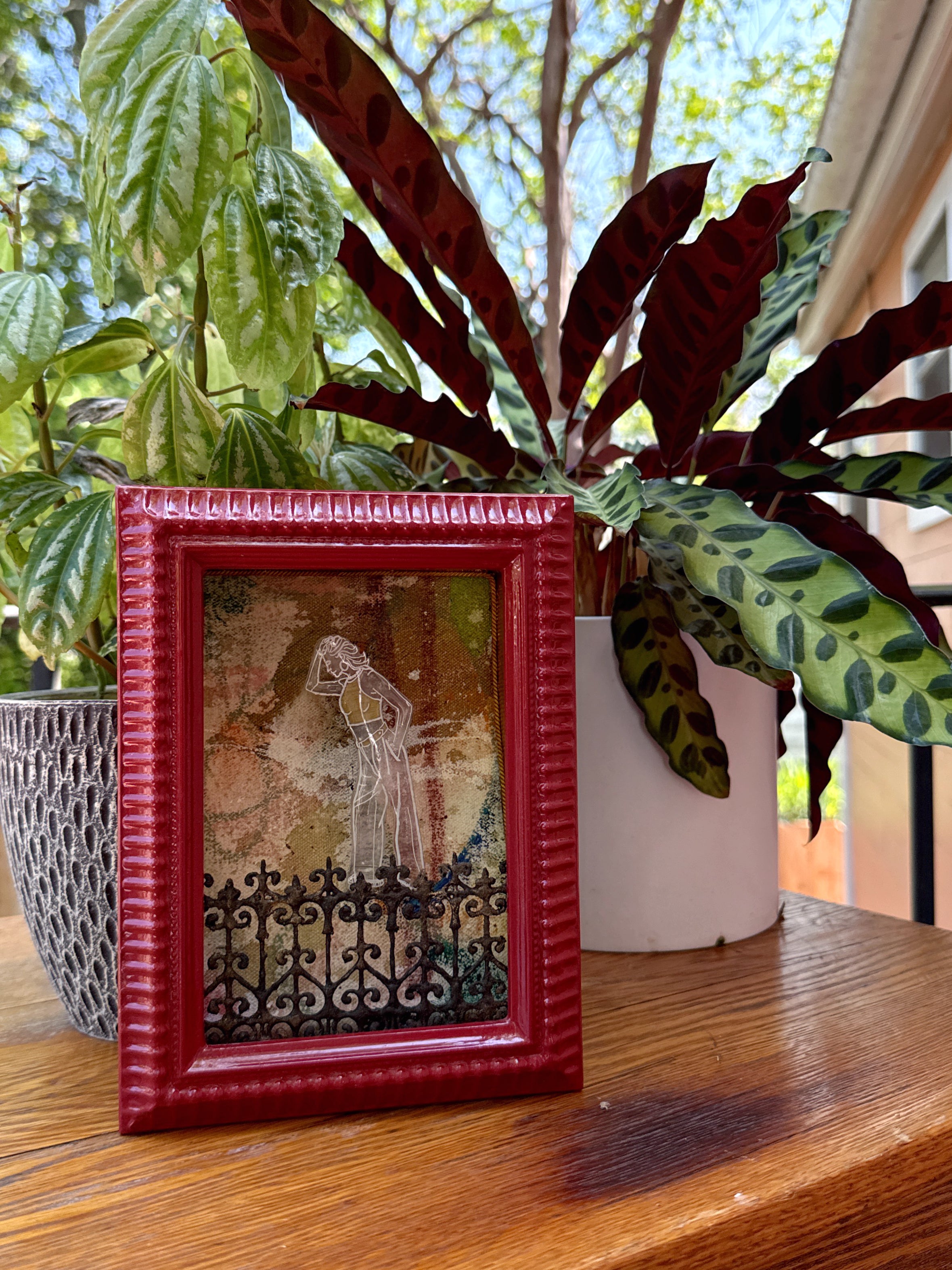Red framed artwork on a wooden surface with Southern plants in the background