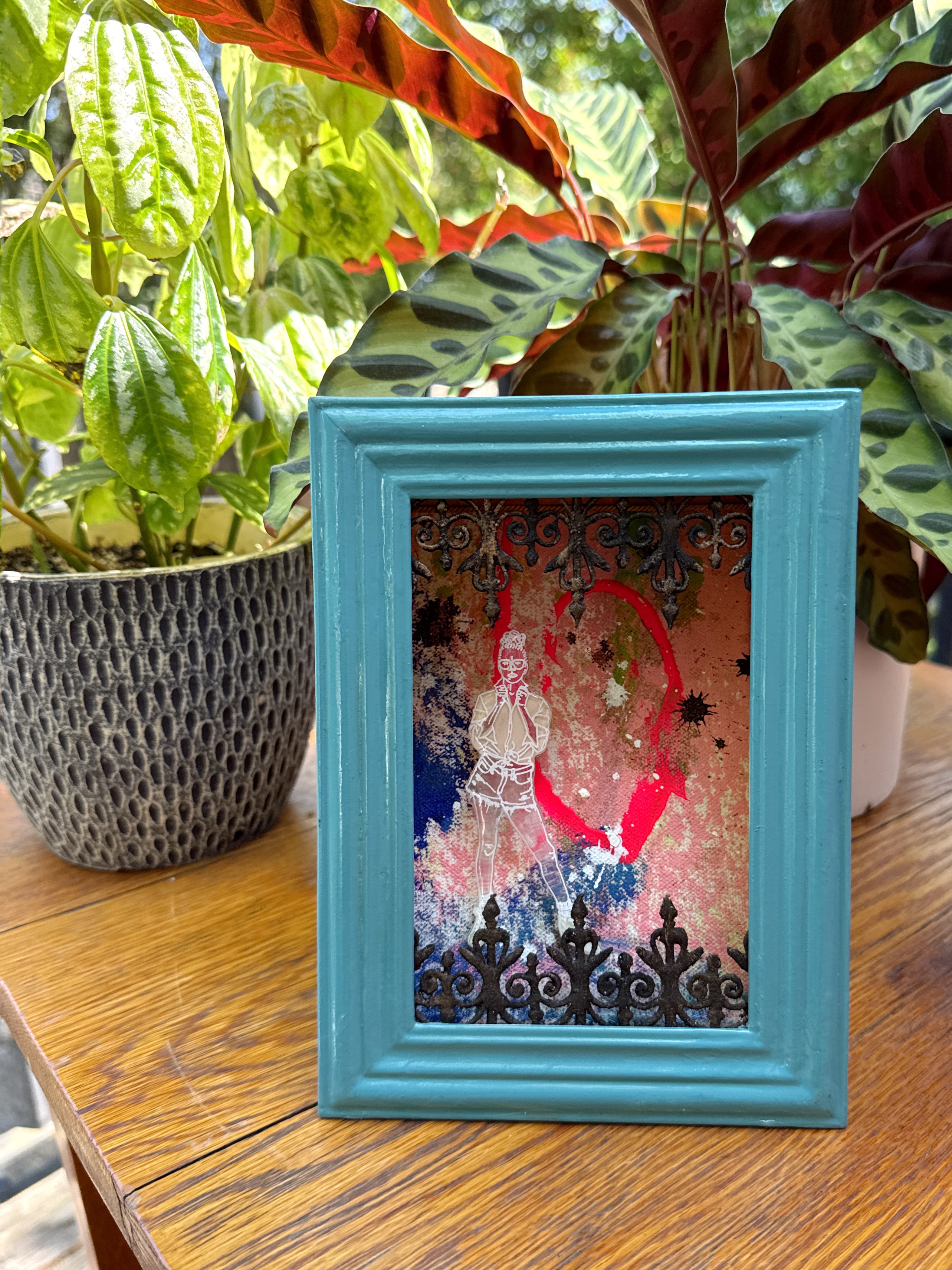Framed artwork with a colorful design on a wooden surface with Southern plants in the background