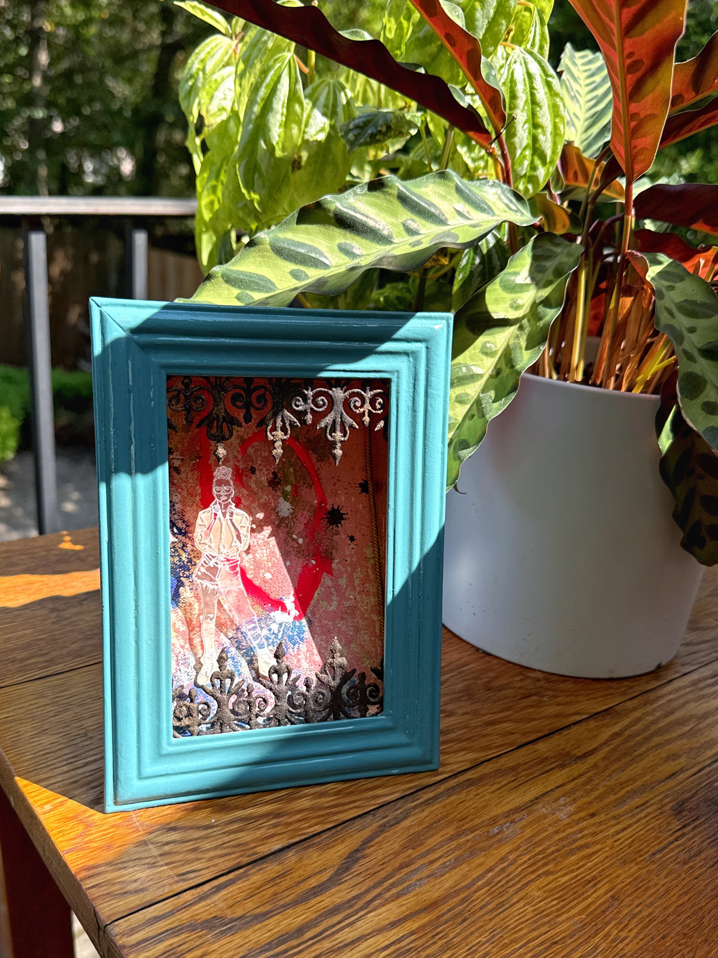 Framed artwork with a colorful figure on a wooden table with southern plants in the background