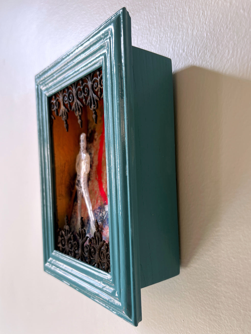 Teal-framed decorative box with a colorful interior design on a white wall.