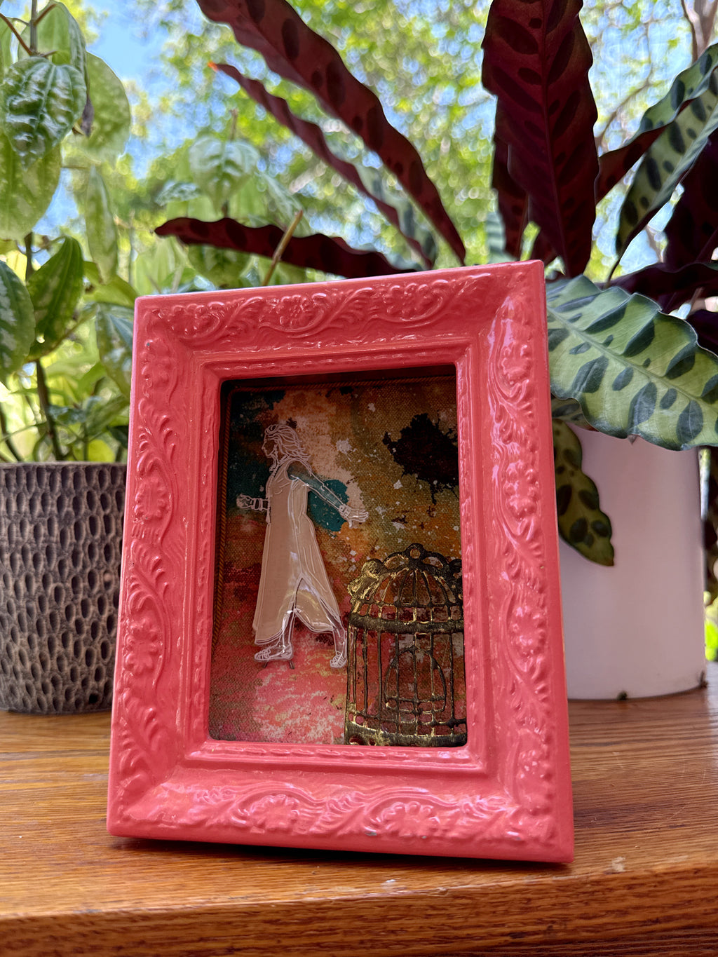 Pink decorative frame with a small artwork on a wooden surface with plants in the background