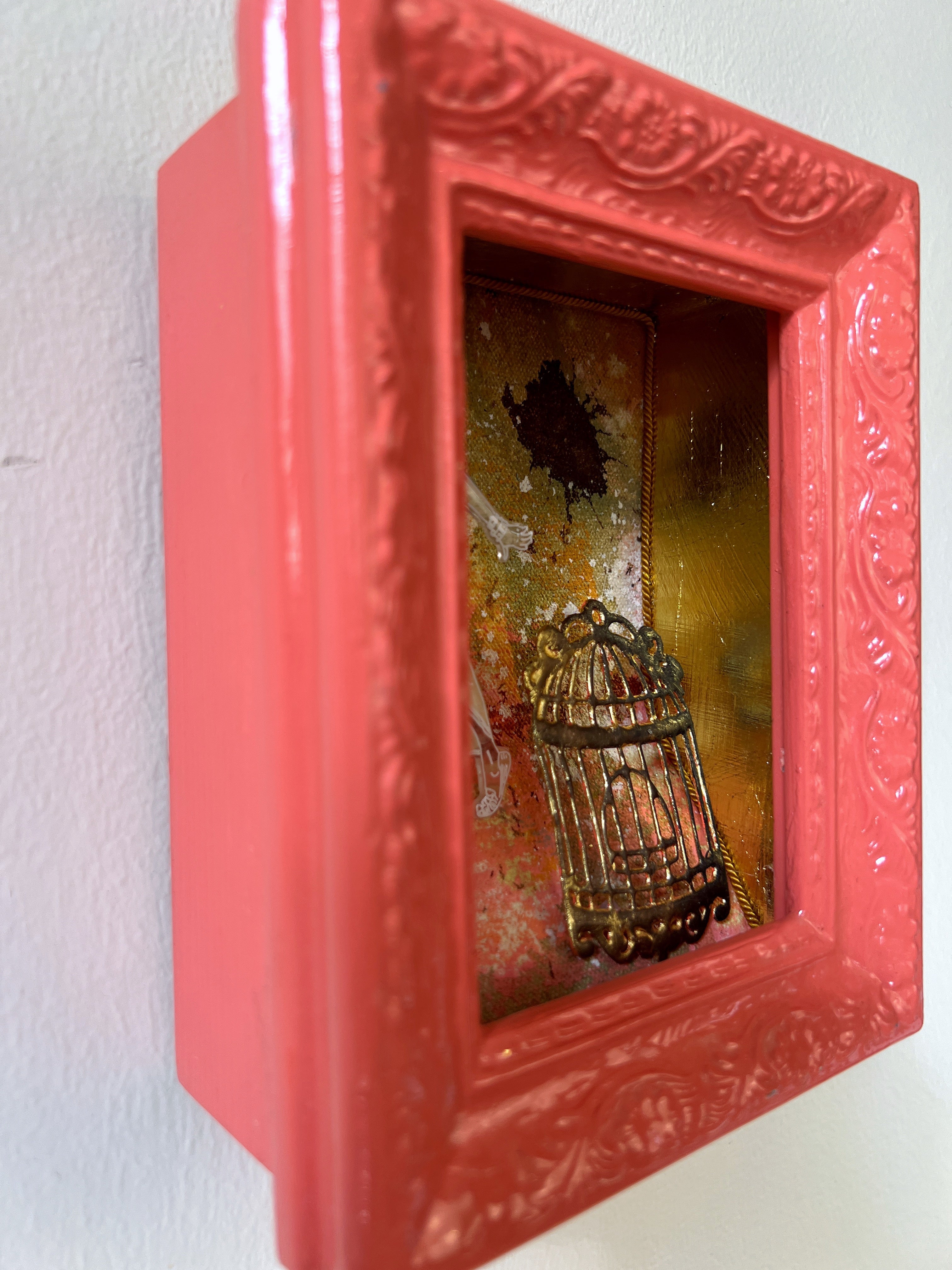 Decorative shadow box with a red frame containing a small birdcage and decorative elements on a white background